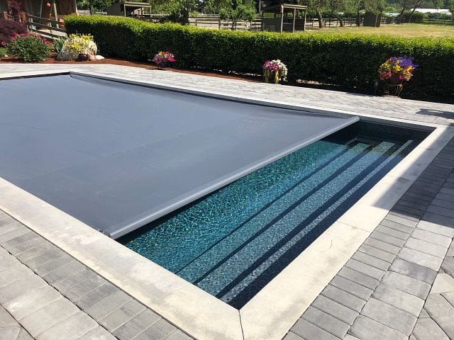 Mostly closed automatic pool cover from Coverstar on a rectangular in-ground fiberglass pool surrounded by a fancy concrete patio and green shrubs.