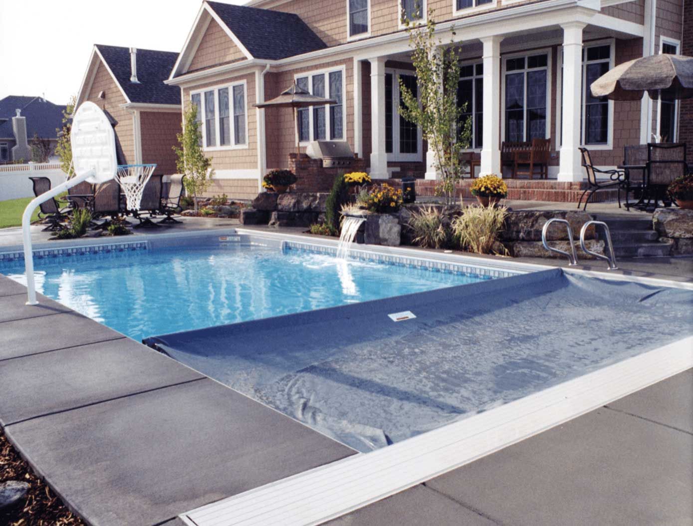 Half open automatic pool cover on a rectangular fiberglass pool in a back yard.