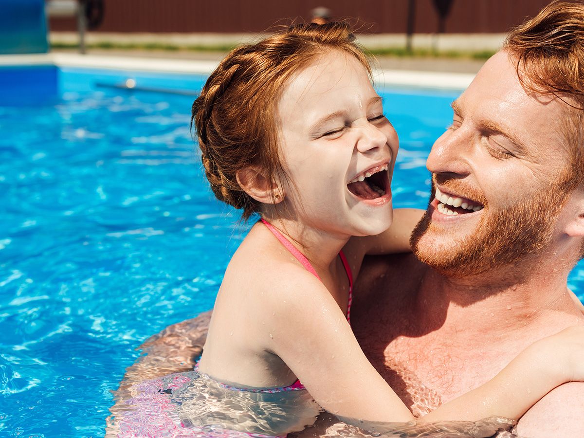 Father holding young daughter and she laughs in a backyard swimming pool.