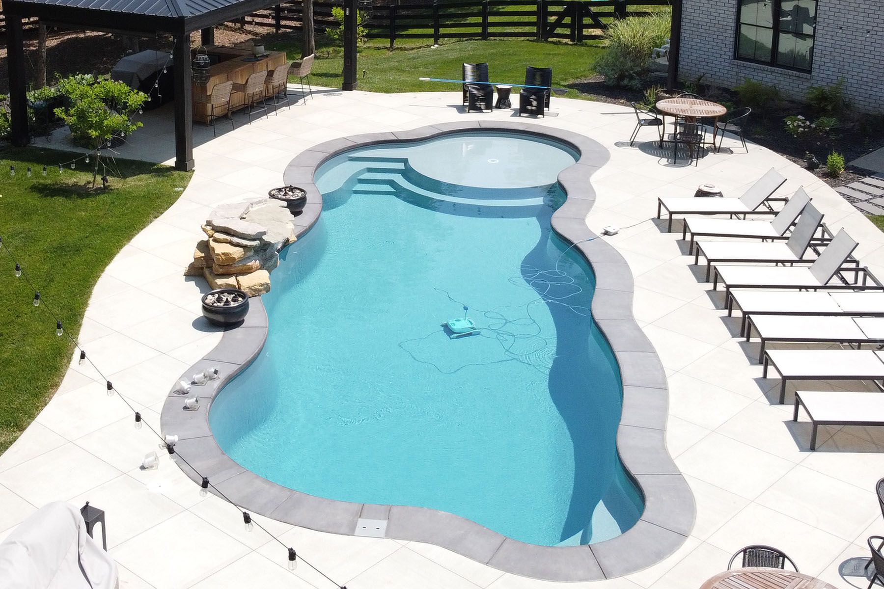 Aerial view of a freeform fiberglass pool with an attratice organic concrete patio in a well-landscaped backyard next to a forest area.