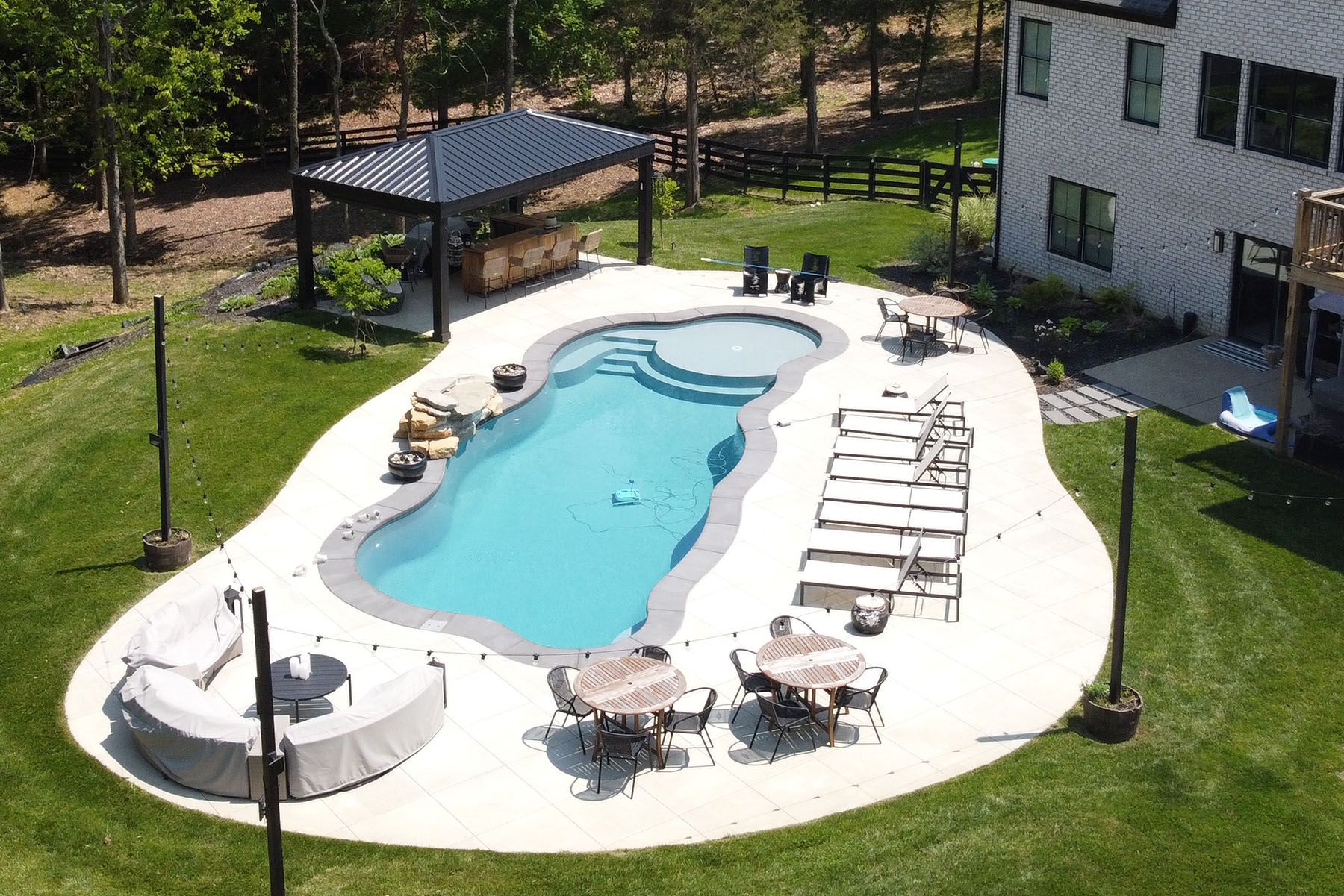 Aerial view of a freeform fiberglass pool with an attratice organic concrete patio in a well-landscaped backyard next to a forest area.