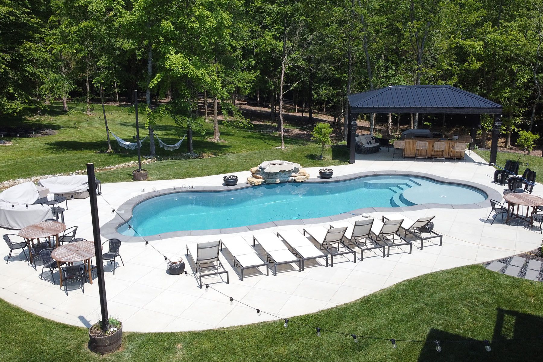 Aerial view of a freeform fiberglass pool with an attratice organic concrete patio in a well-landscaped backyard next to a forest area.