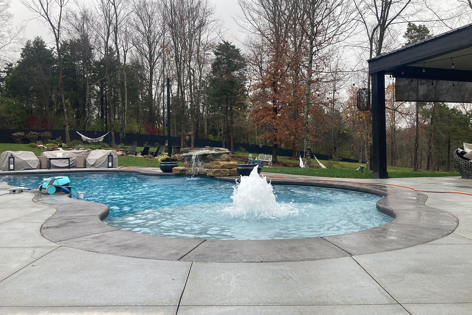 Small water fountain feature in a freeform fiberglass pool with an attratice organic concrete patio in a well-landscaped backyard next to a forest area.