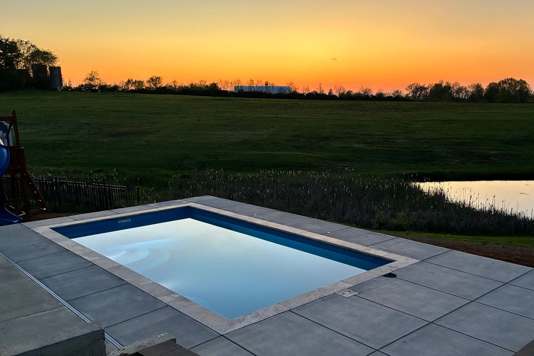 Rectangular in-ground fiberglass swimming pool surrounded by a concrete patio in Shelbyville, Kentucky at sunset.
