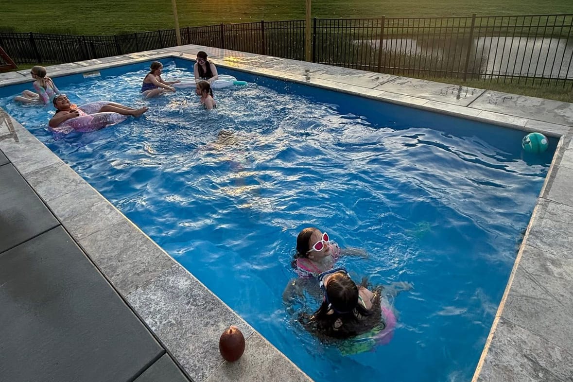 Children playing in a rectangular in-ground fiberglass swimming pool surrounded by a concrete patio in Shelbyville, Kentucky at sunset.