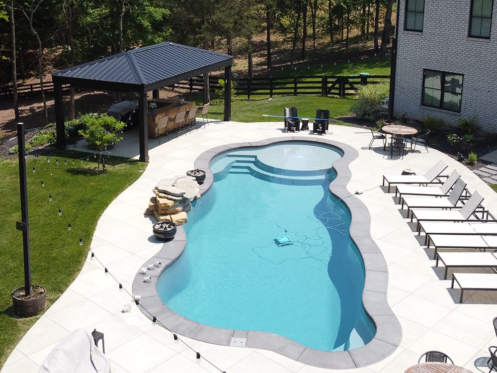 Aerial view of a freeform fiberglass pool with an attratice organic concrete patio in a well-landscaped backyard next to a forest area.