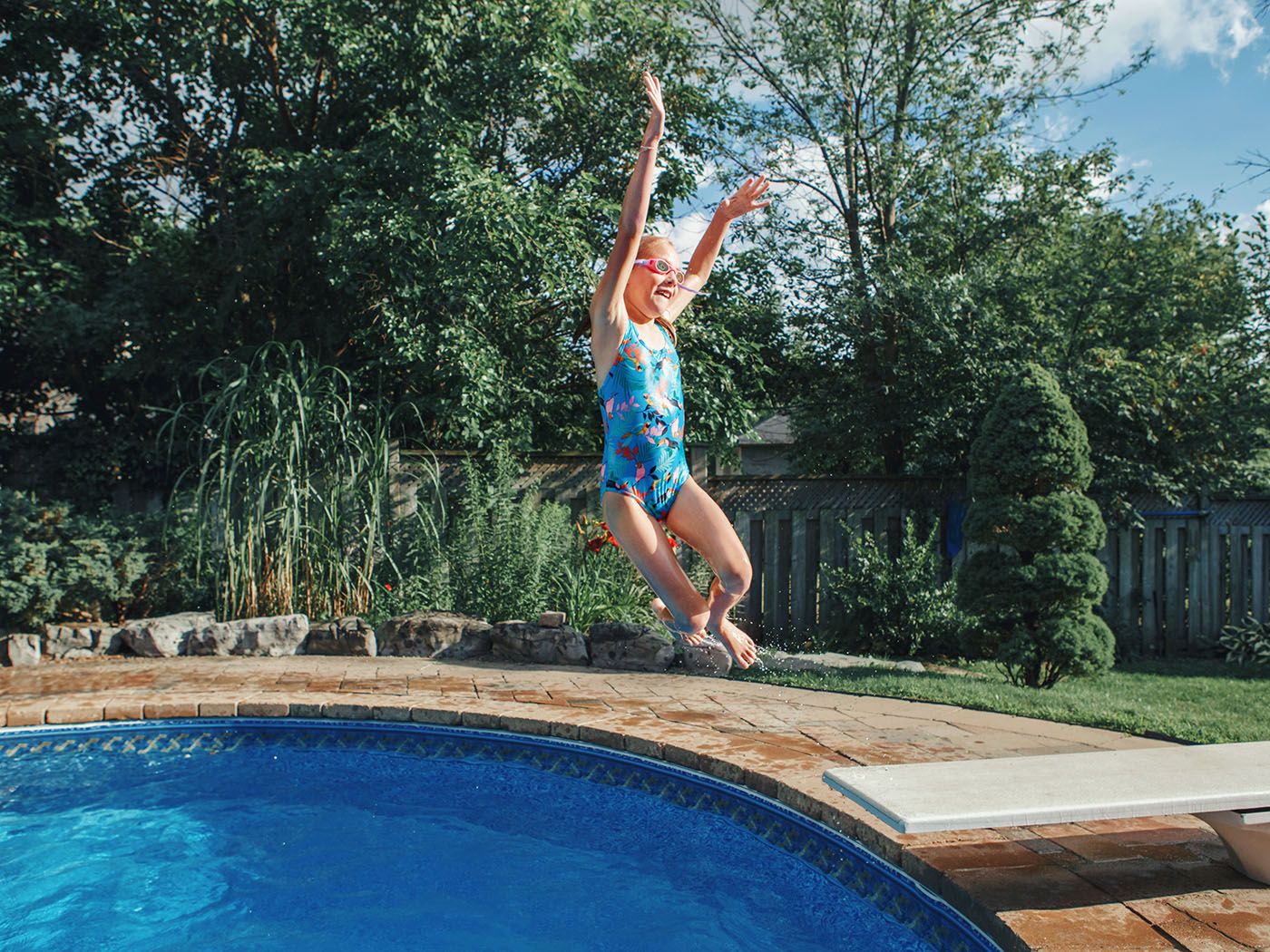 Young girl jumping off of diving board into freeform in-ground fiberglass backyard pool.